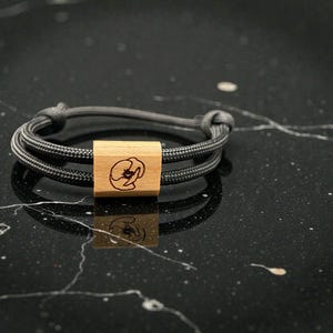 Könnte beinhalten: Ein graues Seilarmband mit einem Mittelstück aus Holz. Das Holzstück ist rechteckig und weist ein geschnitztes Blumendesign auf. Das Armband wird auf einer schwarzen Oberfläche mit weißen Adern gezeigt.