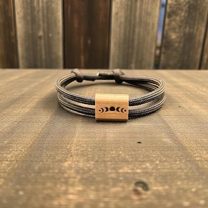Könnte beinhalten: Ein Armband mit einem gewebten, dunkelgrauen Band und einem hellbraunen Mittelstück aus Holz. Das Holzstück ist mit einem Mondphasen-Design graviert. Das Armband wird auf einer Holzoberfläche präsentiert.