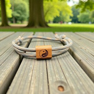 Könnte beinhalten: Ein geflochtenes Armband mit einem Mittelstück aus Holz. Das Armband besteht aus grauem, beigem und blauem Kordel. Das Mittelstück aus Holz zeigt ein Yin-Yang-Symbol. Das Armband liegt auf einer verwitterten Holzoberfläche.