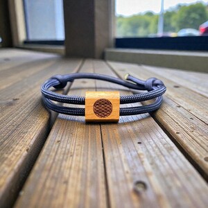 Könnte beinhalten: Ein dunkelblaues Armband mit einem quadratischen Mittelstück aus Holz. Das Quadrat hat ein kreisförmiges Muster eingraviert. Das Armband besteht aus einem gewebten Material und ist an jedem Ende geknotet.