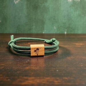 Könnte beinhalten: Ein grünes Seilarmband mit einem quadratischen Holzmittelstück. Das Holzquadrat ist mit einem zarten Löwenzahnmotiv graviert. Das Armband liegt auf einer dunkelbraunen Holzoberfläche.