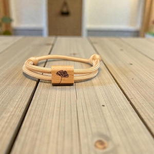 Könnte beinhalten: Ein hellbeige Armband mit einem Mittelstück aus Holz. Das Holzstück ist quadratisch und hat ein dunkles Blumendesign. Das Armband besteht aus mehreren Strängen beigefarbenen Kordels, die an jedem Ende verknotet sind. Das Armband liegt auf einer Holzoberfläche.