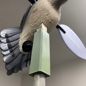 May include: A dove-shaped wind spinner with a light brown body, gray and white wings, and blue eyes. The bird is mounted on a green and silver pole, with a black on/off switch on its side. The wings are white and purple.