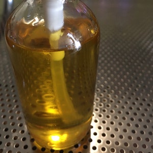 May include: A clear glass bottle with a pump dispenser filled with a light yellow liquid. The bottle is sitting on a silver metal surface with a pattern of small holes.