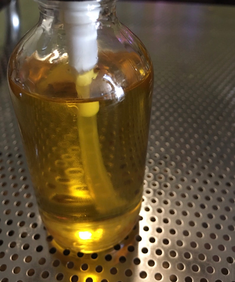 May include: A clear glass bottle with a pump dispenser filled with a light yellow liquid. The bottle is sitting on a silver metal surface with a pattern of small holes.