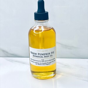 May include: A clear glass bottle with a black dropper top filled with a yellow liquid. The bottle label reads "Warm Pumpkin Pie Botanical Body Oil" and lists the ingredients: Sunflower Oil, Rosehip Oil, Argan Oil, Jojoba Oil, Vitamin E Oil, Fragrance. The bottle is sitting on a white surface.