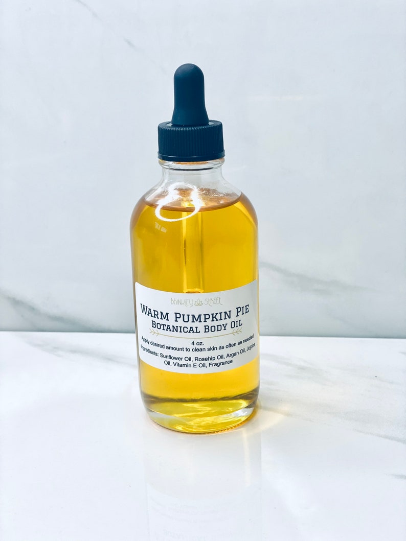 May include: A clear glass bottle with a black dropper top filled with a yellow liquid. The bottle label reads "Warm Pumpkin Pie Botanical Body Oil" and lists the ingredients: Sunflower Oil, Rosehip Oil, Argan Oil, Jojoba Oil, Vitamin E Oil, Fragrance. The bottle is sitting on a white surface.