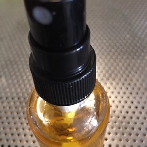 May include: A clear glass bottle with a black spray nozzle filled with a yellow liquid. The bottle is sitting on a silver surface with a pattern of small holes.