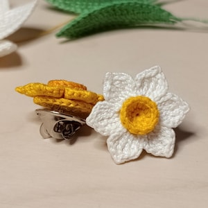 Puede incluir: Un broche de flor de ganchillo con seis pétalos blancos y un centro amarillo elevado. La parte posterior del broche muestra un mecanismo de alfiler de metal plateado. La flor está hecha con hilo fino, mostrando una apariencia texturizada.