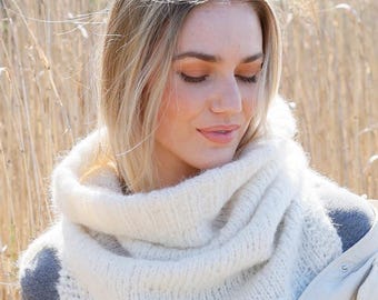 May include: A close-up of a woman wearing a cream-colored, chunky knit cowl neck scarf. The scarf is wrapped around the neck and shoulders. The woman is wearing a gray top and a light-colored jacket. The background is blurred.