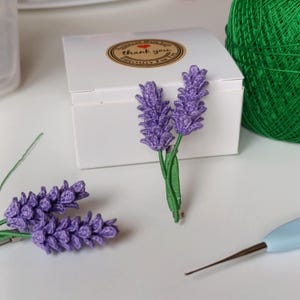 May include: Handmade lavender flower brooches. The brooches are purple with green stems. One brooch is pinned to a green ribbon. A white box with a "thank you" sticker is in the background. A ball of green yarn and a crochet hook are also visible.