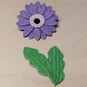 Op de afbeelding: Een handgemaakte gehaakte bloem in paarse tinten, met een wit en zwart centrum. Er zijn ook twee groene gehaakte bladeren aanwezig. De bloem heeft gedetailleerd bloemblaadjeswerk en de bladeren hebben een getextureerde uitstraling. De items zijn waarschijnlijk voor knutselen of decoratie.
