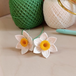 Puede incluir: Dos flores de narciso de ganchillo hechas a mano con pétalos blancos y centros amarillos. Las flores se colocan frente a ovillos de hilo verdes y blancos. Un ganchillo es visible en el fondo.