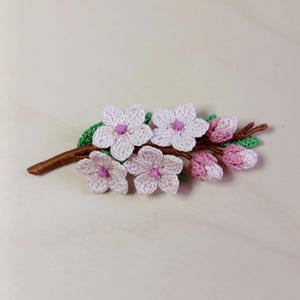 May include: A crocheted floral branch with five white flowers with pink centres, pink buds, and green leaves. The branch is brown. The handmade craft is set against a light-coloured background.