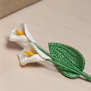 Puede incluir: Dos flores de cala blancas tejidas a ganchillo con centros amarillos y una hoja verde. Las flores hechas a mano están dispuestas sobre una superficie clara. Los delicados detalles del ganchillo son visibles, mostrando la textura y la artesanía del diseño floral.