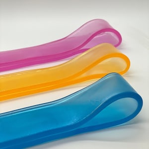 May include: Three translucent rubber bands in pink, orange, and blue. The bands are arranged in a stack, with the pink band on top, the orange band in the middle, and the blue band on the bottom.