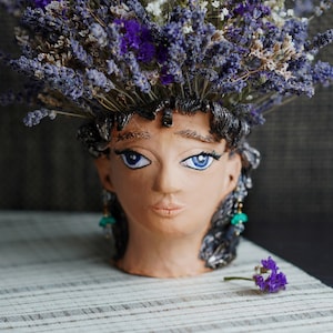 Puede incluir: Un macetero de cerámica con forma de cabeza de mujer con ojos azules y flores de lavanda en el pelo. El macetero está sobre una superficie rayada blanca y marrón.