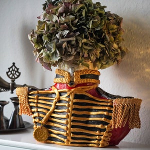 Op de afbeelding: Een decoratieve vaas in de vorm van een militair uniform met gouden en zwarte versieringen. De vaas is gevuld met gedroogde hortensia's.
