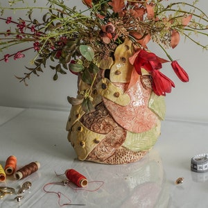 Könnte beinhalten: Eine Keramikvase mit einem strukturierten, mehrfarbigen Design, gefüllt mit einem Strauß roter und rosa Blumen. Die Vase steht auf einer weißen Oberfläche mit Garnrollen, einer Schere und einem Maßband.