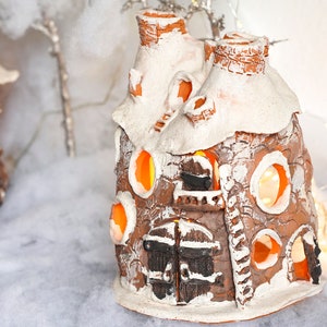 May include: Two small, brown, ceramic houses with white snow-like accents and glowing orange lights inside. The houses are sitting on a white, fluffy surface.