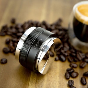 May include: A black and silver coffee tamper with the word "CREMA" engraved on the side. The tamper is surrounded by coffee beans.