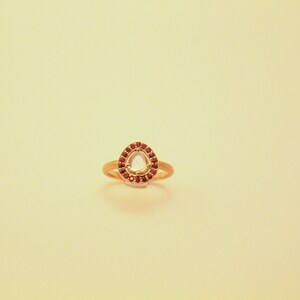May include: A gold ring with a pear-shaped clear gemstone surrounded by a halo of small red gemstones.