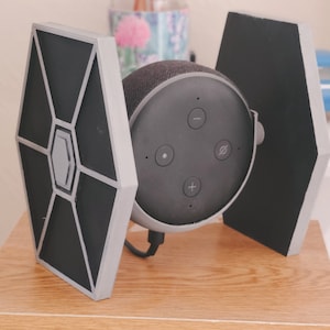 May include: A black and grey Amazon Echo Dot smart speaker with a custom 3D printed TIE fighter stand. The stand is made of white plastic and has a black TIE fighter design.