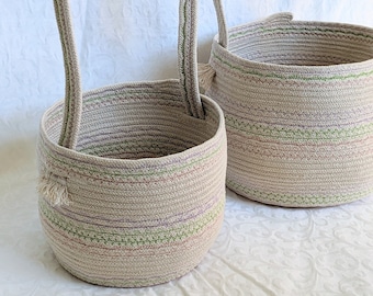 May include: Two woven rope baskets with handles. The baskets are beige with green and purple stripes. The smaller basket is in front of the larger basket.