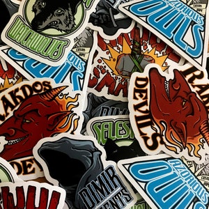 May include: A collection of colourful stickers featuring various designs, including a wolf, a devil, a creature with wings, and a skull. The stickers are printed on white paper and have a glossy finish.