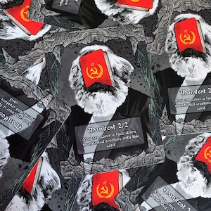 Puede incluir: Cartas con una ilustración en blanco y negro de una figura barbuda con un libro rojo con un martillo y una hoz dorados. Las cartas tienen el texto "Manifest 2/2" y "Puedes cubrir una criatura manifestada boca abajo con esta carta."
