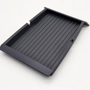 May include: A rectangular, black plastic tray with a ribbed interior. The tray has a raised border and is designed with a recessed area. The object is set against a white background.