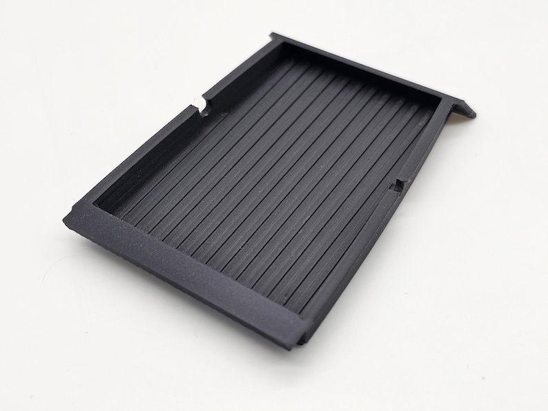 May include: A rectangular, black plastic tray with a ribbed interior. The tray has a raised border and is designed with a recessed area. The object is set against a white background.