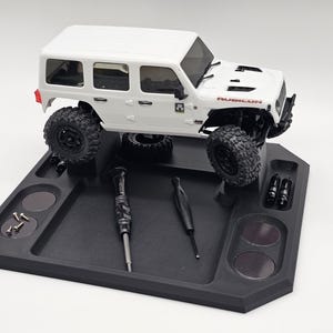 May include: A white toy Jeep Wrangler Rubicon model sits on a black tray with compartments. The tray holds tools and small parts, including screws and what appear to be shock absorbers. The Jeep has black wheels and the word "RUBICON" is visible.