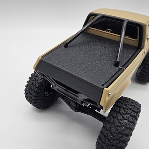 May include: Tan and black toy pickup truck with large, textured tires. The truck features a black roll bar and a textured black bed cover. The vehicle is set against a white background.