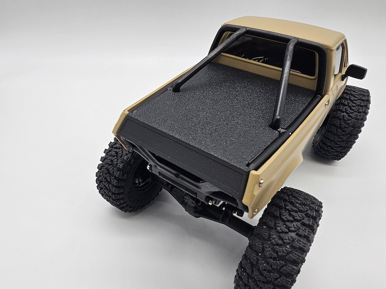 May include: Tan and black toy pickup truck with large, textured tires. The truck features a black roll bar and a textured black bed cover. The vehicle is set against a white background.