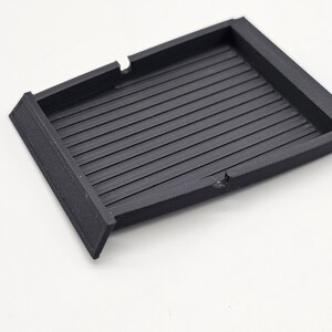 May include: A black, rectangular tray with a ridged interior. The tray has a raised border and two small cutouts on one side. The object is made of a matte material and sits on a white surface.