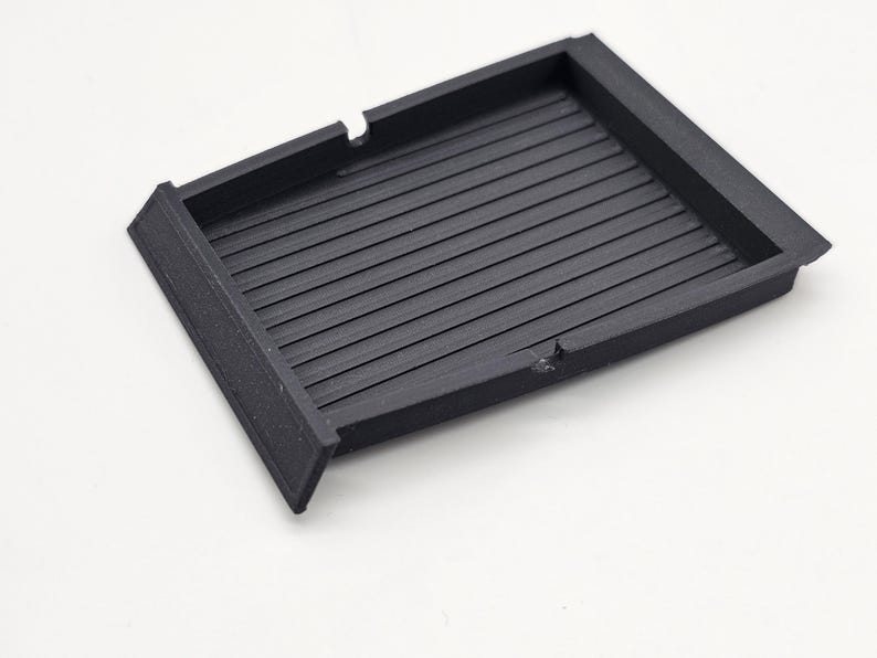 May include: A black, rectangular tray with a ridged interior. The tray has a raised border and two small cutouts on one side. The object is made of a matte material and sits on a white surface.