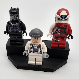May include: Three Lego minifigures on a black display stand. The minifigures are a black superhero with a purple chest, a white minifigure with a gray hat and a black and gray torso, and a red minifigure with a white helmet and a red and black torso.