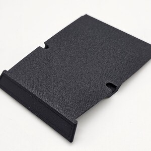 May include: A black, rectangular plastic component with a textured surface. It features a raised edge on one side and two curved cutouts. The object is designed for a specific purpose, likely as part of a larger assembly.