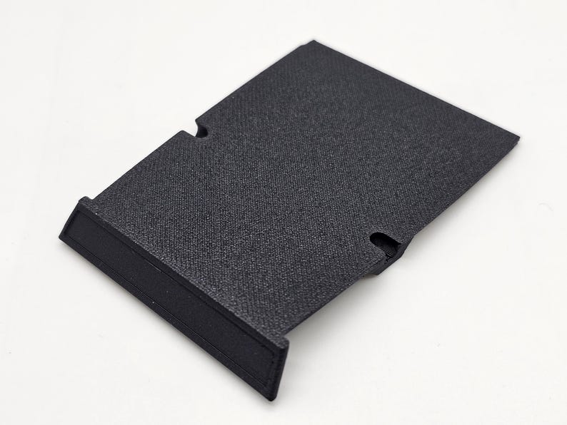 May include: A black, rectangular plastic component with a textured surface. It features a raised edge on one side and two curved cutouts. The object is designed for a specific purpose, likely as part of a larger assembly.