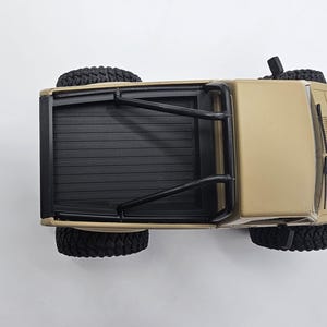 May include: Tan-colored toy pickup truck with black tires and a black roll bar. The truck bed is black with horizontal grooves. The tires are large and have a rugged tread pattern. The vehicle is viewed from above.
