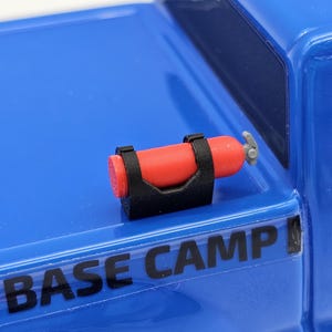 May include: A miniature red fire extinguisher with a gray nozzle, secured by a black bracket, is mounted on a blue surface. The text "BASE CAMP" is visible on the blue surface. The extinguisher is a small-scale model.