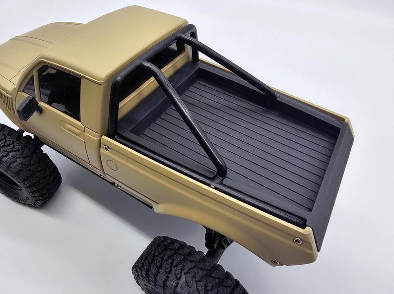 May include: Tan-colored toy pickup truck with black roll bar and black bed. The truck has large, black, off-road tires. The truck's cab has a window and side mirror. The bed has a textured surface.