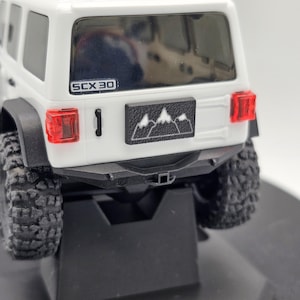 May include: A white toy SUV with black tires and a black bumper. The rear of the vehicle features red taillights, a black license plate with a mountain graphic, and the text "SCX 30".