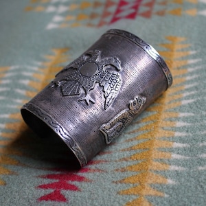 May include: A silver cuff bracelet with an intricate design of two eagles with their wings spread. The bracelet is textured and has a raised design.