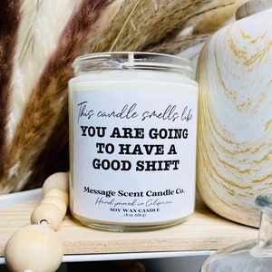 May include: A white soy wax candle with a black label that reads "This candle smells like YOU ARE GOING TO HAVE A GOOD SHIFT". The label also includes the text "Message Scent Candle Co. Hand poured in California (8 oz. 226 g)".