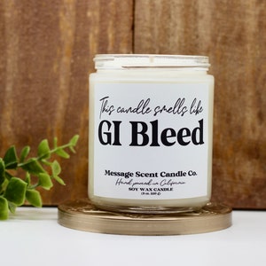 May include: A white soy wax candle in a clear glass jar with a black label that reads "This candle smells like GI Bleed". The label also includes the text "Message Scent Candle Co. Hand poured in California. Soy Wax Candle (8 oz / 226 g)".