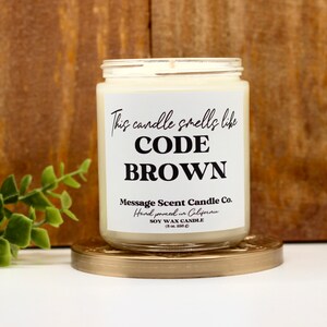 May include: A white soy wax candle with a black label that reads "This candle smells like CODE BROWN" and "Message Scent Candle Co. Hand poured in California".