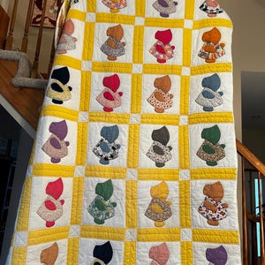 May include: A handmade quilt featuring a repeating pattern of doll-like figures in various colors. The quilt has a white background with yellow borders and a grid-like design. The dolls wear dresses with floral patterns.