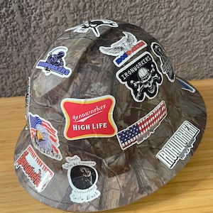 May include: A camouflage hard hat with various stickers, including "Ironworker High Life", "Ironworkers", and "Death Calls For Everyone".